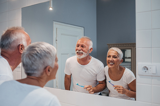 Elderly couple smiling and brushing their teeth in the bathroom mirror.
