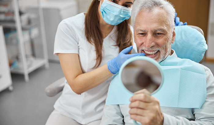 Older man sitting in a dental chair looking in a handheld mirror at his new dental implants from Fusion Dental Specialists in Happy Valley, OR